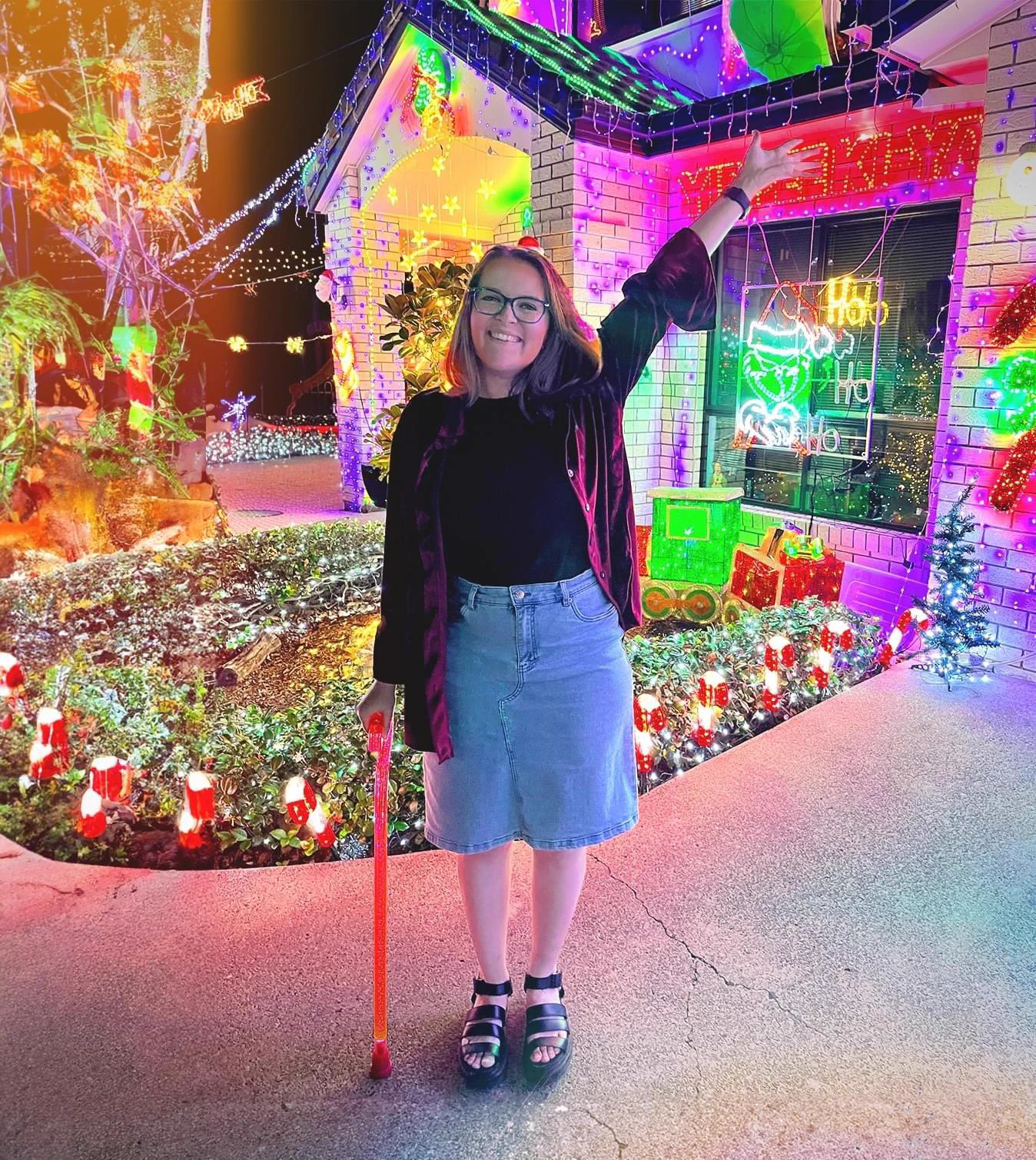 Photo of a white woman with short brown hair and blue eyes. She wears blue glasses, a maroon velvet jacket, and a denim skirt. She is standing in front of a Christmas light display while holding a bright pink walking stick. She is smiling at the camera.