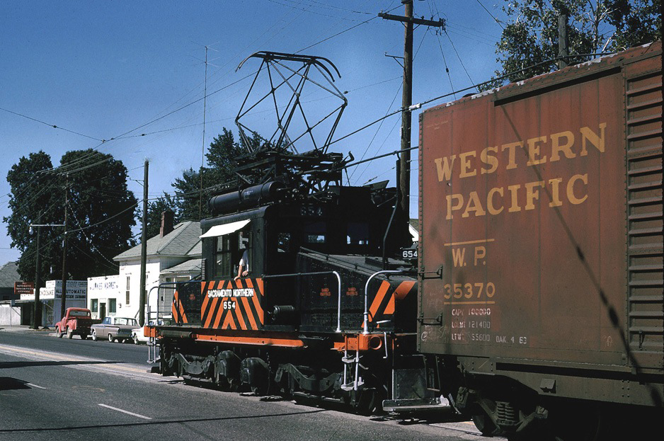 Sacramento Northern steeple-cab electric #654 has a cut of boxcars from the Del Monte plant at Yuba City, CA as it makes its way down Bridge Street on August 4, 1964. The electrified operations ended here the following year but the trackage remained in use until 2004. Drew