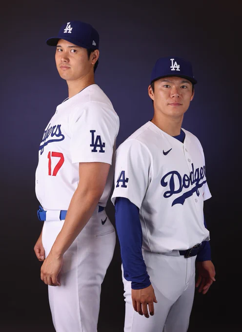 Shohei Ohtani and Yoshinobu Yamamoto stand back-to-back for a posed photo. They wear white Los Angeles Dodgers uniforms and blue Dodgers caps.
