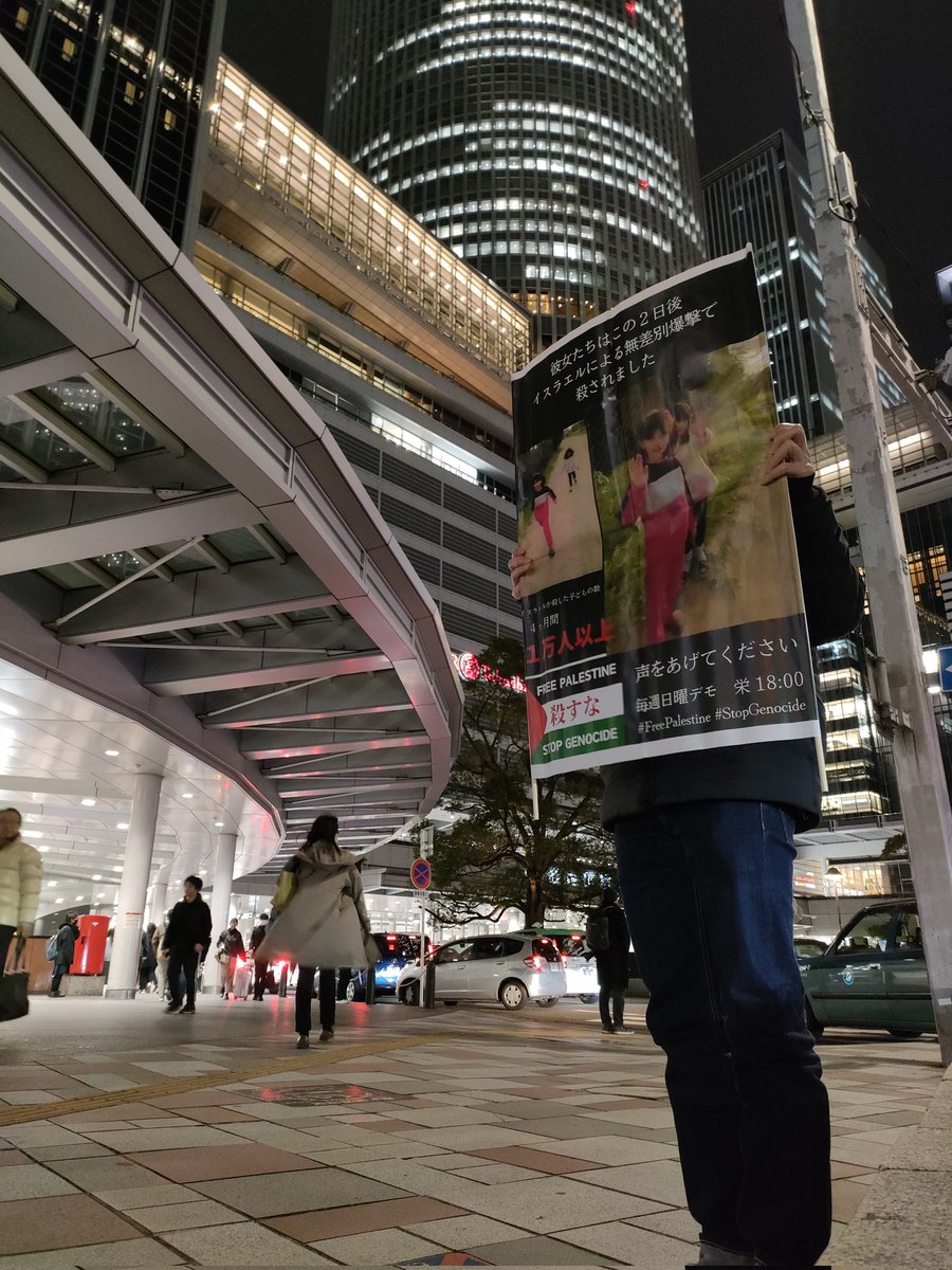 プラスターを掲げ路上で一人立っている。
プラスターには２人の少女の写真と「彼女たちはこの２日後　イスラエルによる無差別爆撃で　殺されました」「声をあげてください　毎週日曜デモ　栄１８：００」の文字。