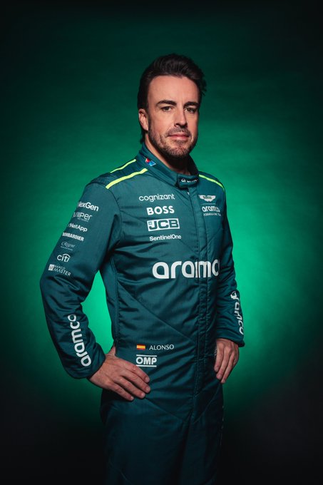 A photo of Fernando Alonso posing with his hands on his hips and staring at the camera, wearing his 2024 race suit 