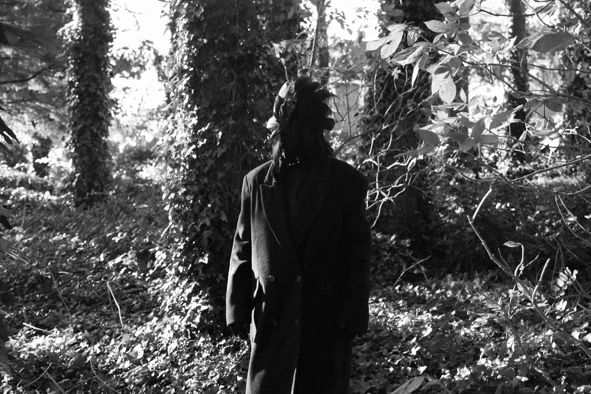 Black and white photograph of a dark figure wearing a large coat, standing in an overgrown forest.
