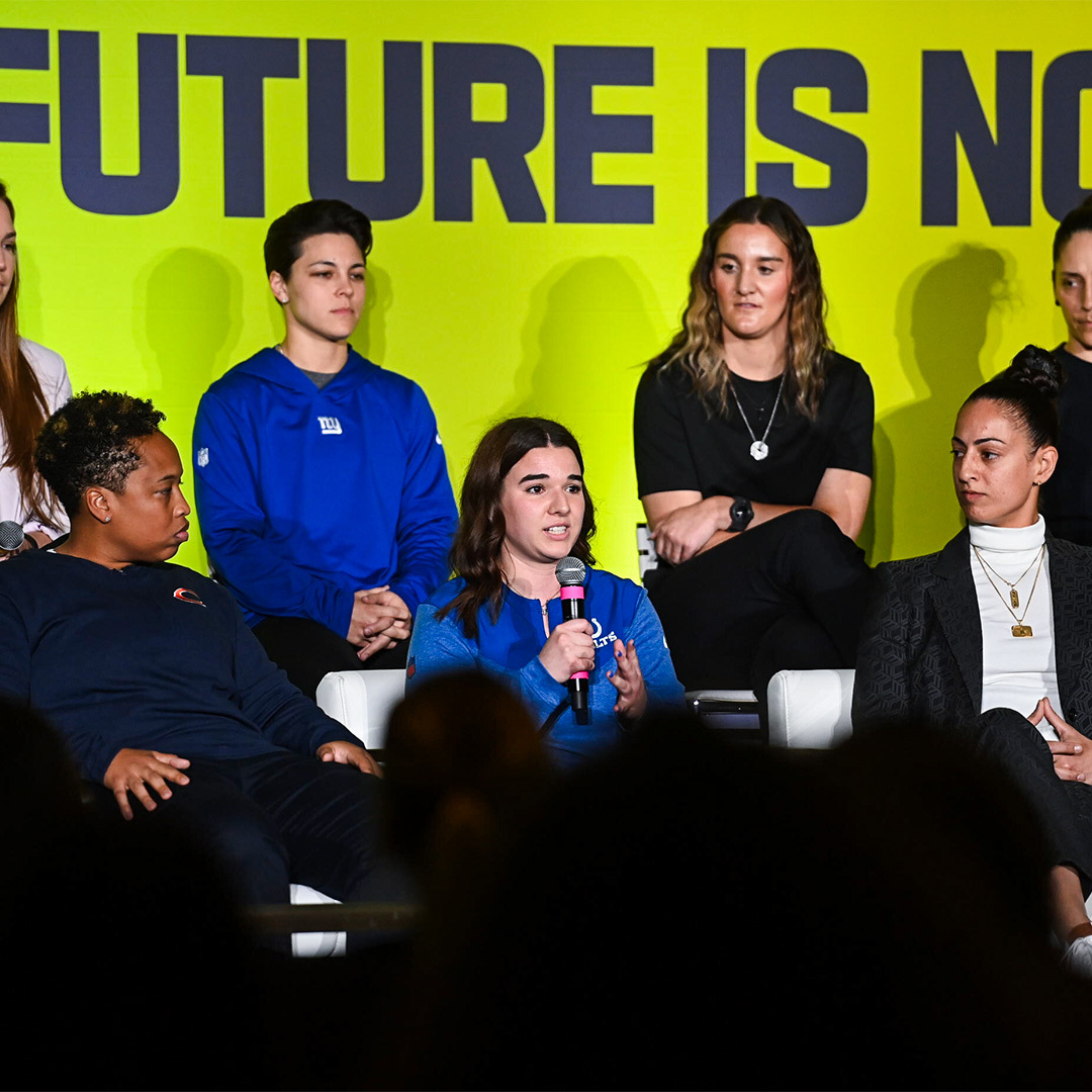 Izzy Diaz speaks on the NFL's Women in Football panel at the 2024 NFL Combine. She is seated around other women who are participating in the discussion and is holding the mic. Behind the women there is a backdrop that reads "The future is now".