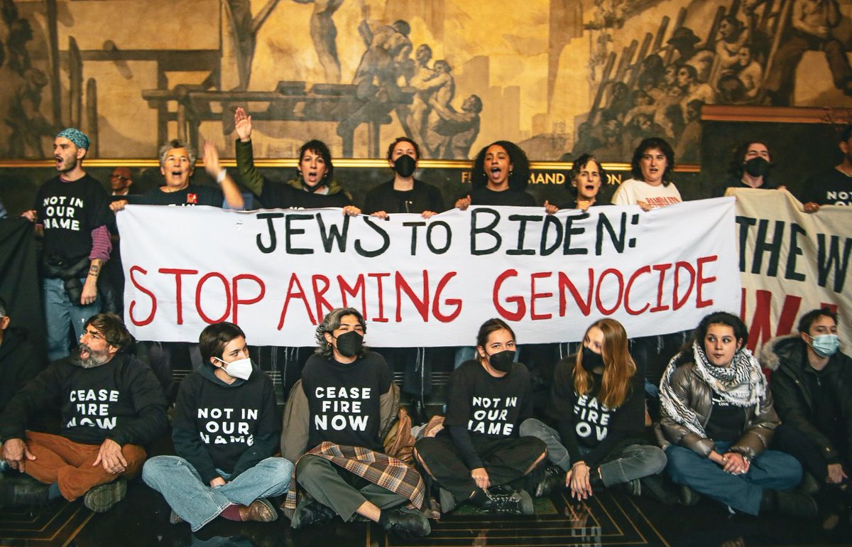 group of mostly masked protesters in lobby of 30 Rock over Biden TV appearance. sign reads "JEWS TO BIDEN: STOP ARMING GENOCIDE". seats read "CEASEFIRE NOW"; "NOT IN OUR NAME."