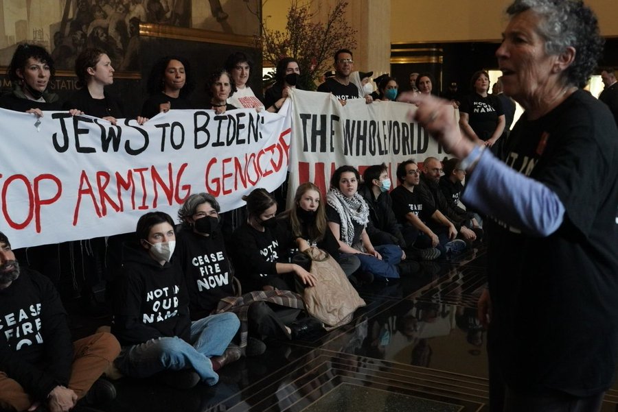 standing protester speaks to group of mostly masked protesters in lobby of 30 Rock over Biden TV appearance. sign reads "JEWS TO BIDEN: STOP ARMING GENOCIDE". seats read "CEASEFIRE NOW"; "NOT IN OUR NAME."