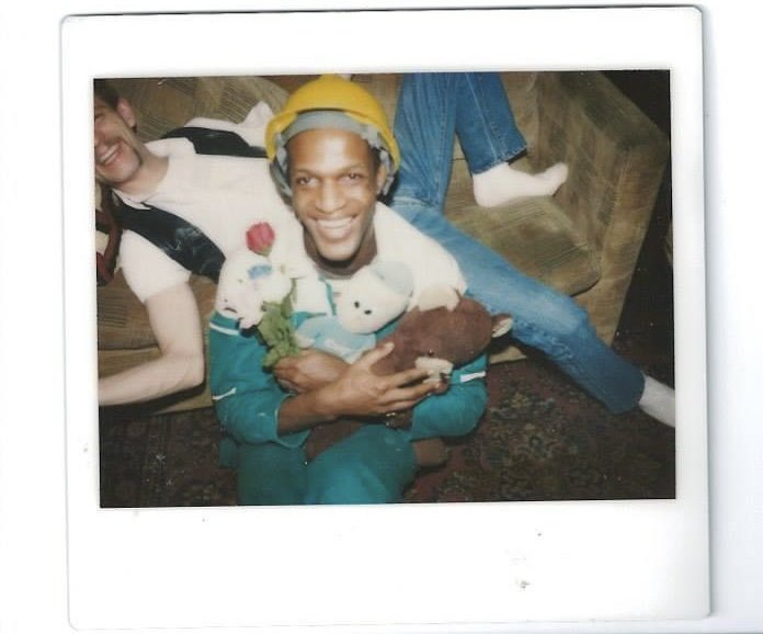 Rare photo of iconic black trans activist Marsha P. Johnson, holding flowers and a teddy bear, captured in a vibrant moment. She wears a yellow hard hat and chic blue and white attire, her radiant smile illuminating the frame. Photographer and photo date unknown.📸