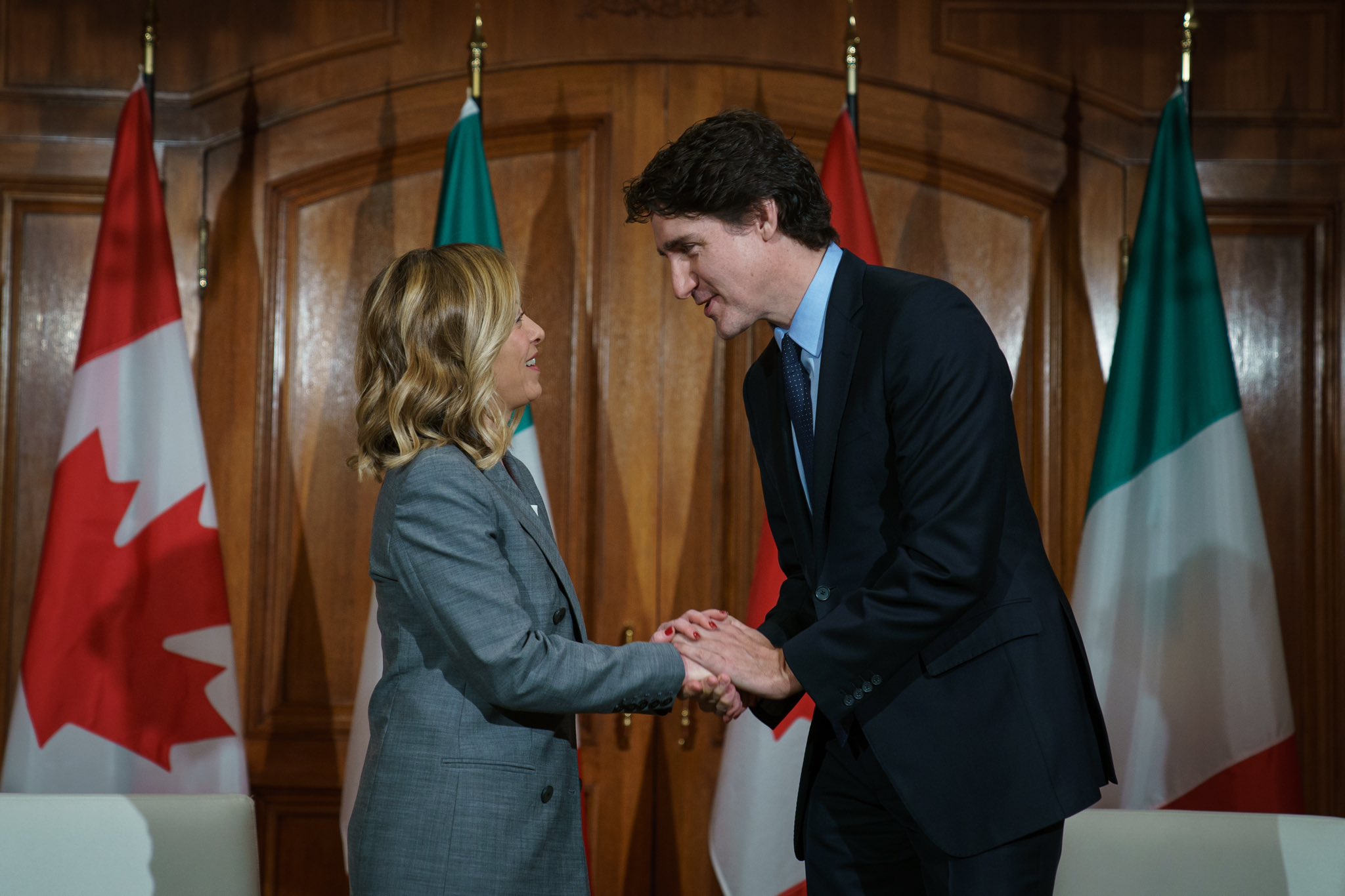 Prime Minister Justin Trudeau and Prime Minister Giorgia Meloni are standing and shaking hands. Prime Minister Trudeau is speaking and Prime Minister Meloni is smiling. Two Canadian flags and two Italian flags are displayed behind them.