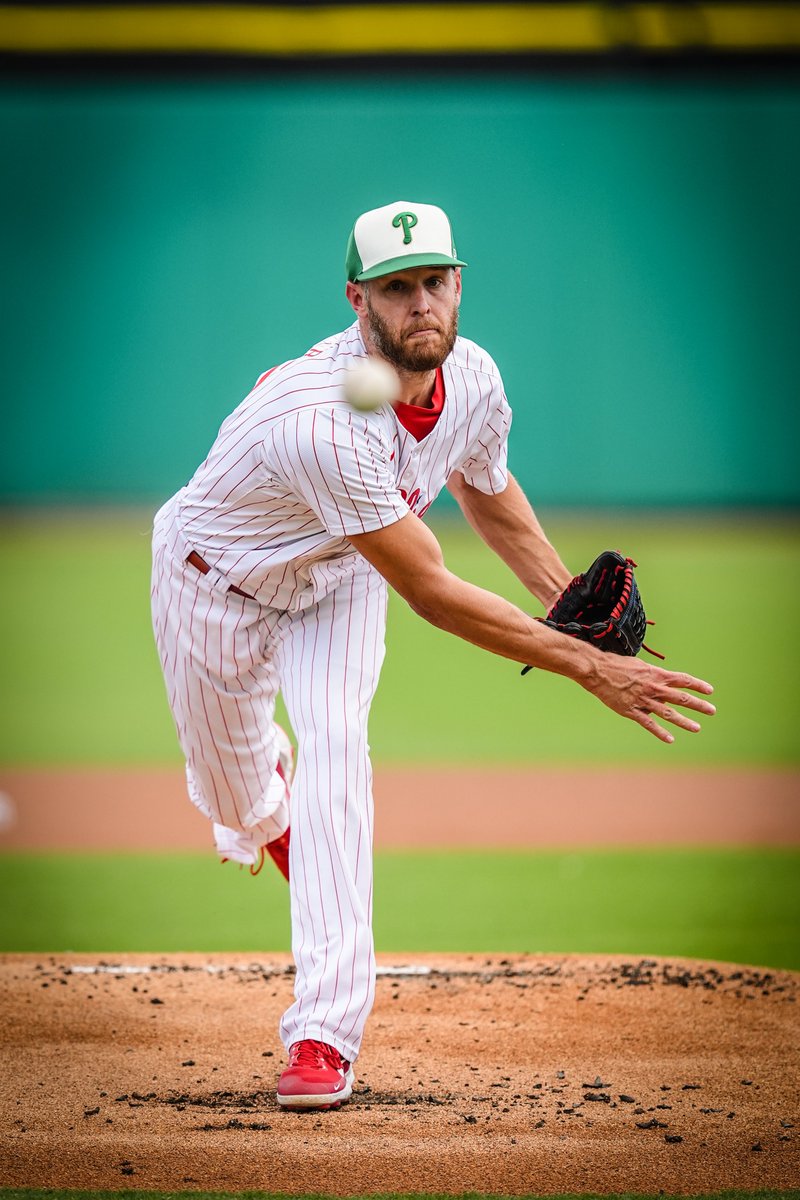 Zack Wheeler throwing a pitch while the ball he threw heads toward home plate. He's wearing the red pinstripes uniform and a white and green Phillies hat for St. Patrick's Day.