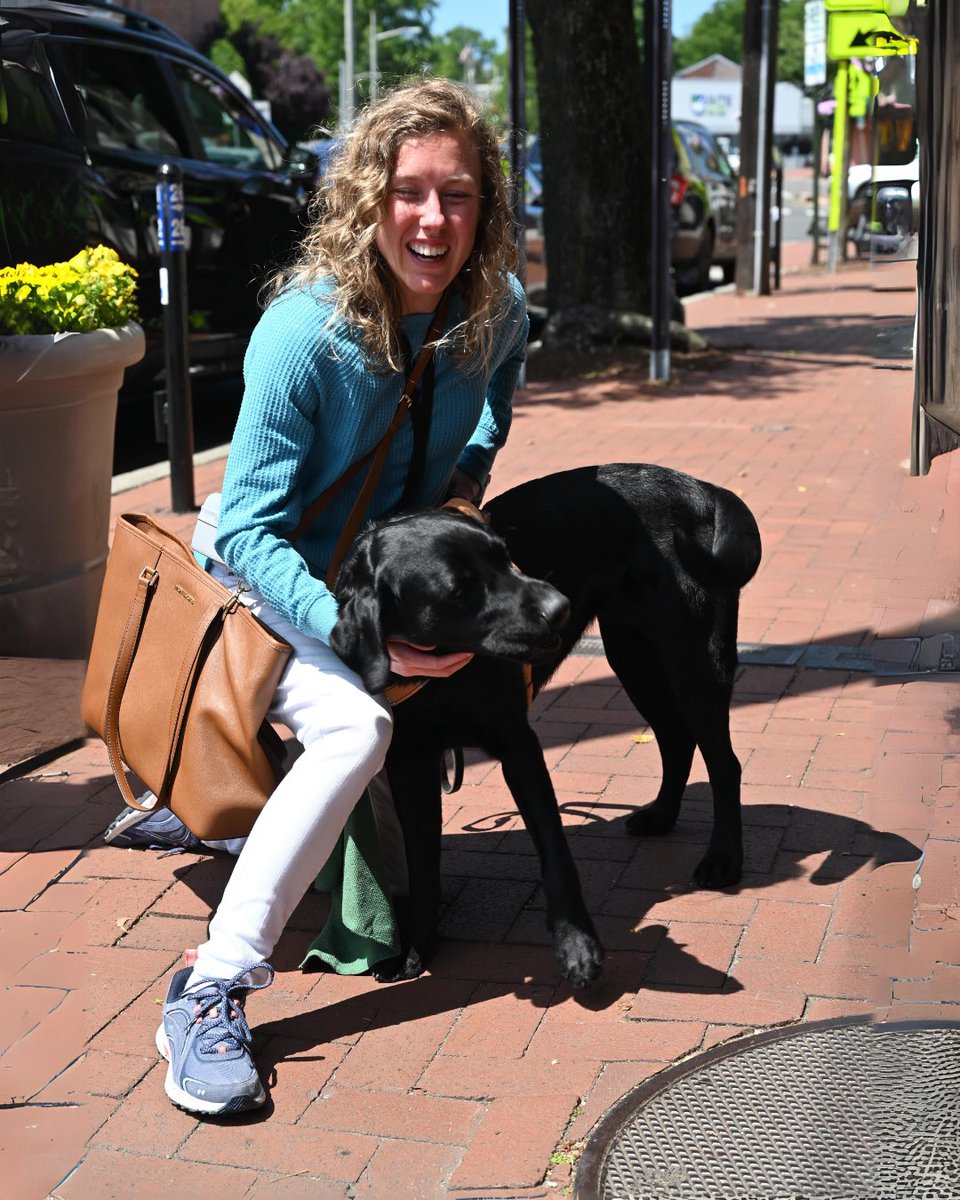 Warmer days are almost here ☀😎 Although we may have lost an hour of sleep, the good news is that the sun will set past 7 p.m. starting this week 🌆 Image shows a woman kneeling next to her black guide dog laughing. Her guide dog is leaning into her while she pets its back.