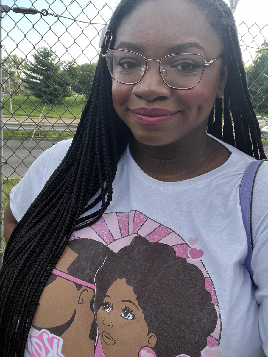 A photo of myself standing outside in front of a fence. I’m smiling and wearing a shirt displaying a Black version of Barbie and Ken.