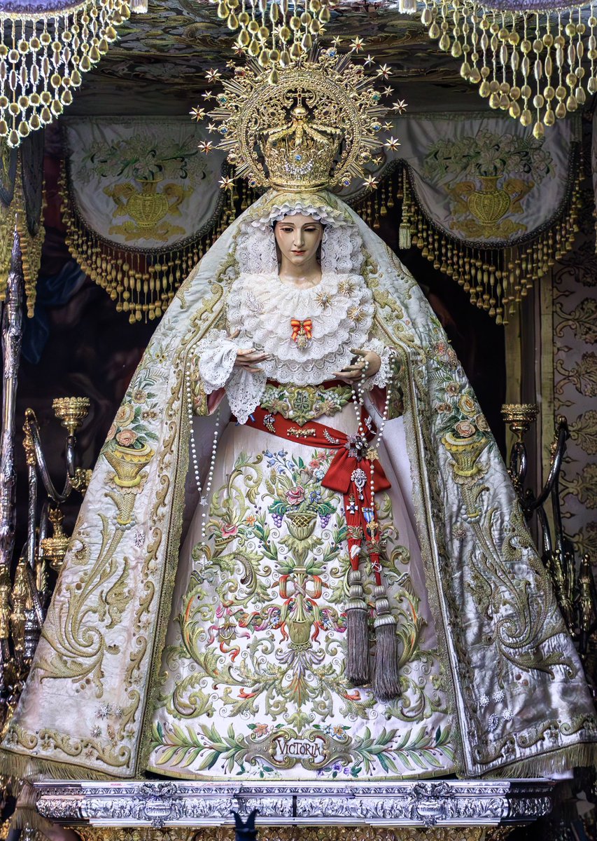 María Santísima de la Victoria se presenta vestida de Reina por su vestidor, Francisco Garví,auxiliado por la Camarera Mayor y la Camarista de la Virgen. Lleva saya de salida, de tisú de plata bordada en oro y sedas, el manto de salida de damasco igualmente bordado en oro y sedas