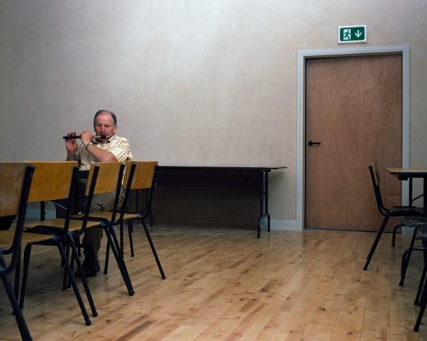 A man sits alone at a communal meeting table, playing the flute. The walls are bare and sparse. He looks as if he is remembering more than just the tune he’s playing. #BookReview of Brian Newman's photobook 'Association' by Colin Graham @19acres. Read on arinsproject.com