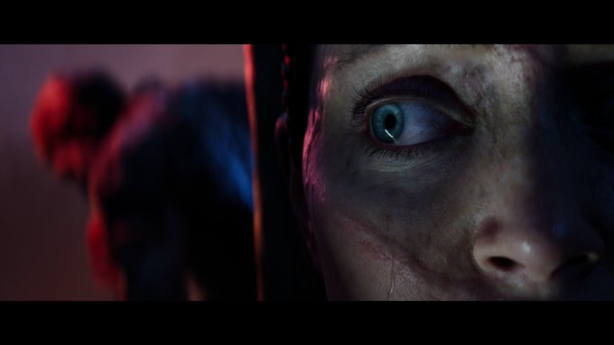 A close up of Senua’s face rendered in incredible detail. Out of focus in the background a figure bathed in red light turns to her direction.