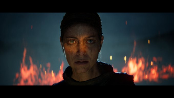 Senua looks into the camera, her face in shadow except for a glow of orange light across her eyes. Flames rise behind her.