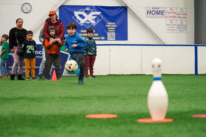 On the indoor soccer field, a student kicks a soccer ball toward a bowling pin as others look on