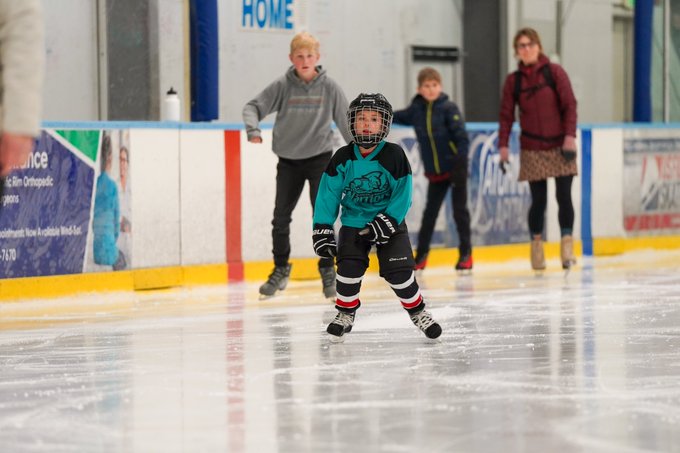 A young ice skater in hockey gear skates on ice rink during event while others look on and skate in background