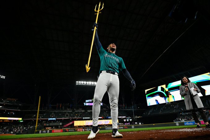 Julio Rodríguez yells and stands on the field after getting a walk-off hit while holding up a gold trident. He wears a teal jersey with silver lettering.