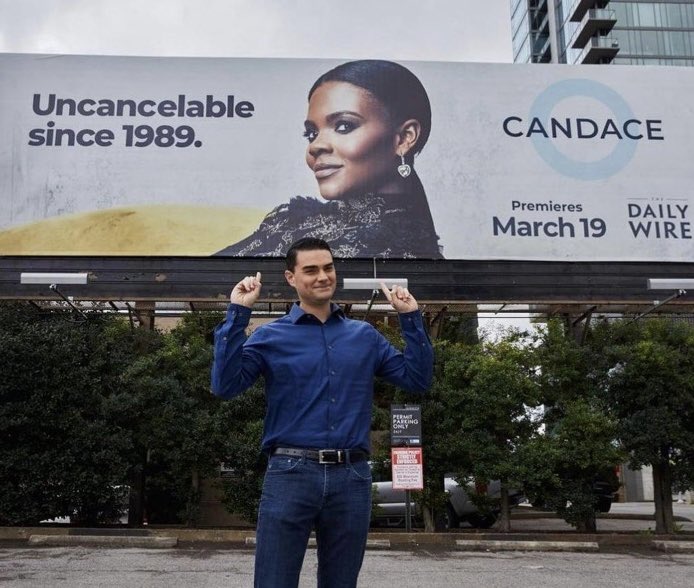 A photo of Ben Shapiro standing in-font of a Candace Owens billboard that says “Uncancelable since 1989” taken in 2021