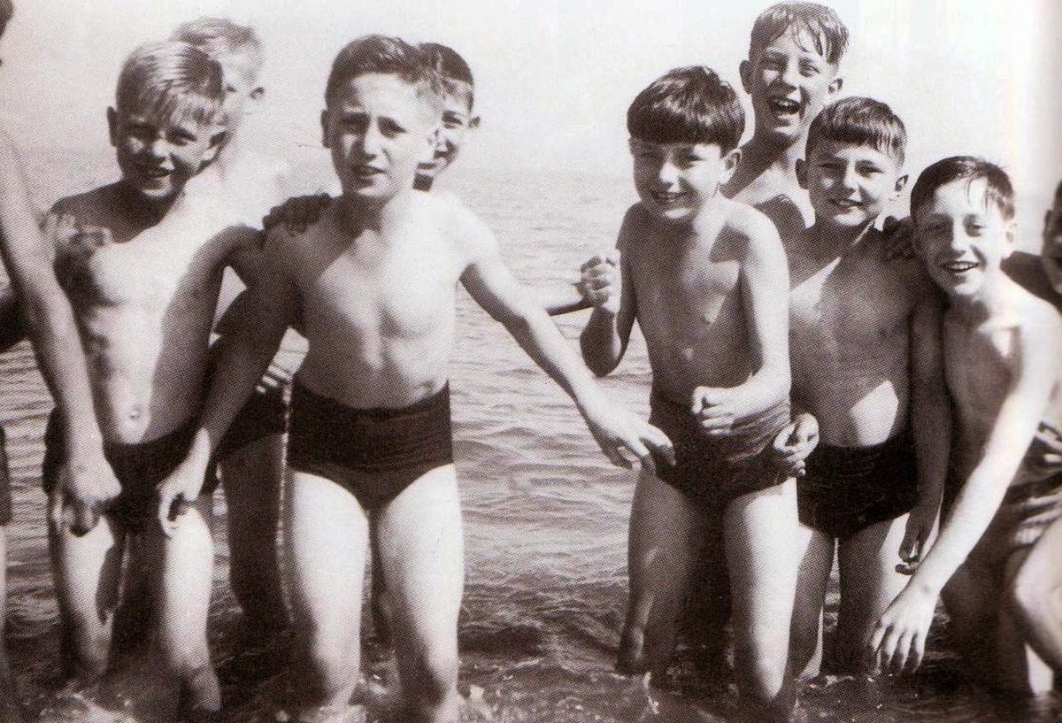 1951, Isle of Man. Pupils from Dovedale Junior School, Liverpool on a school trip. Peter Sissons (newsreader & journalist) has a hand on the shoulder of pal John Lennon (musician) who is next to James Tarbuck ('comedian') who is in front of Brian Labone (footballer)