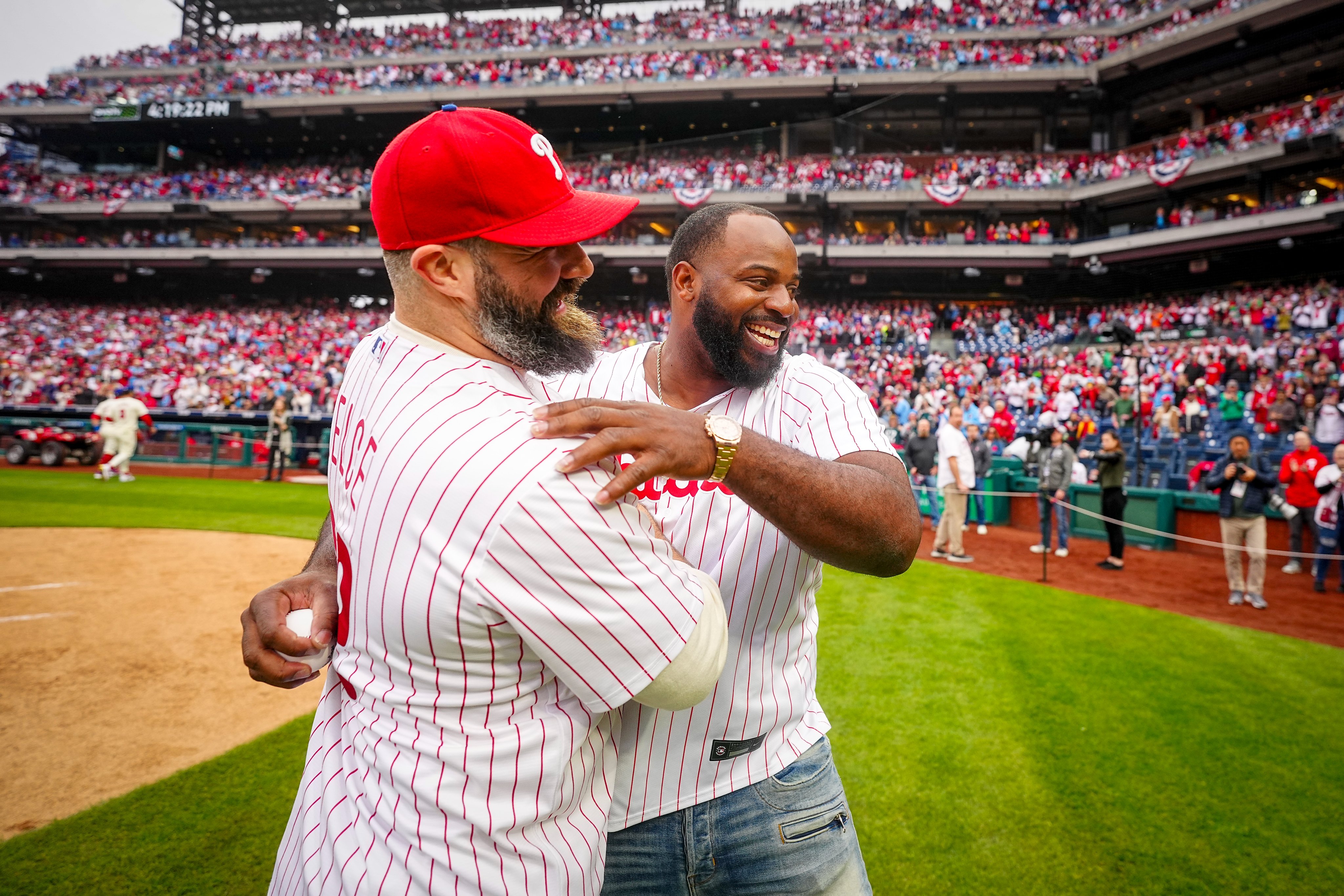 Jason Kelce and Fletcher Cox hugging each other after throwing out the first pitch. They're both wearing red pinstripes Phillies jerseys.
