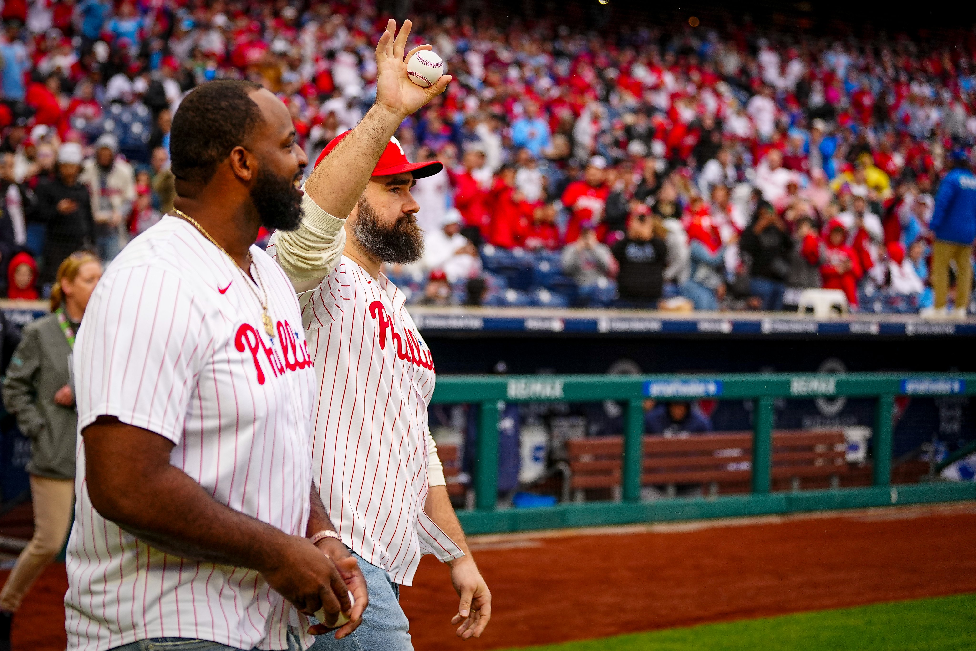 Jason Kelce and Fletcher Cox walking onto the field to throw out the first pitch before the game. They're both wearing red pinstripes Phillies jerseys.