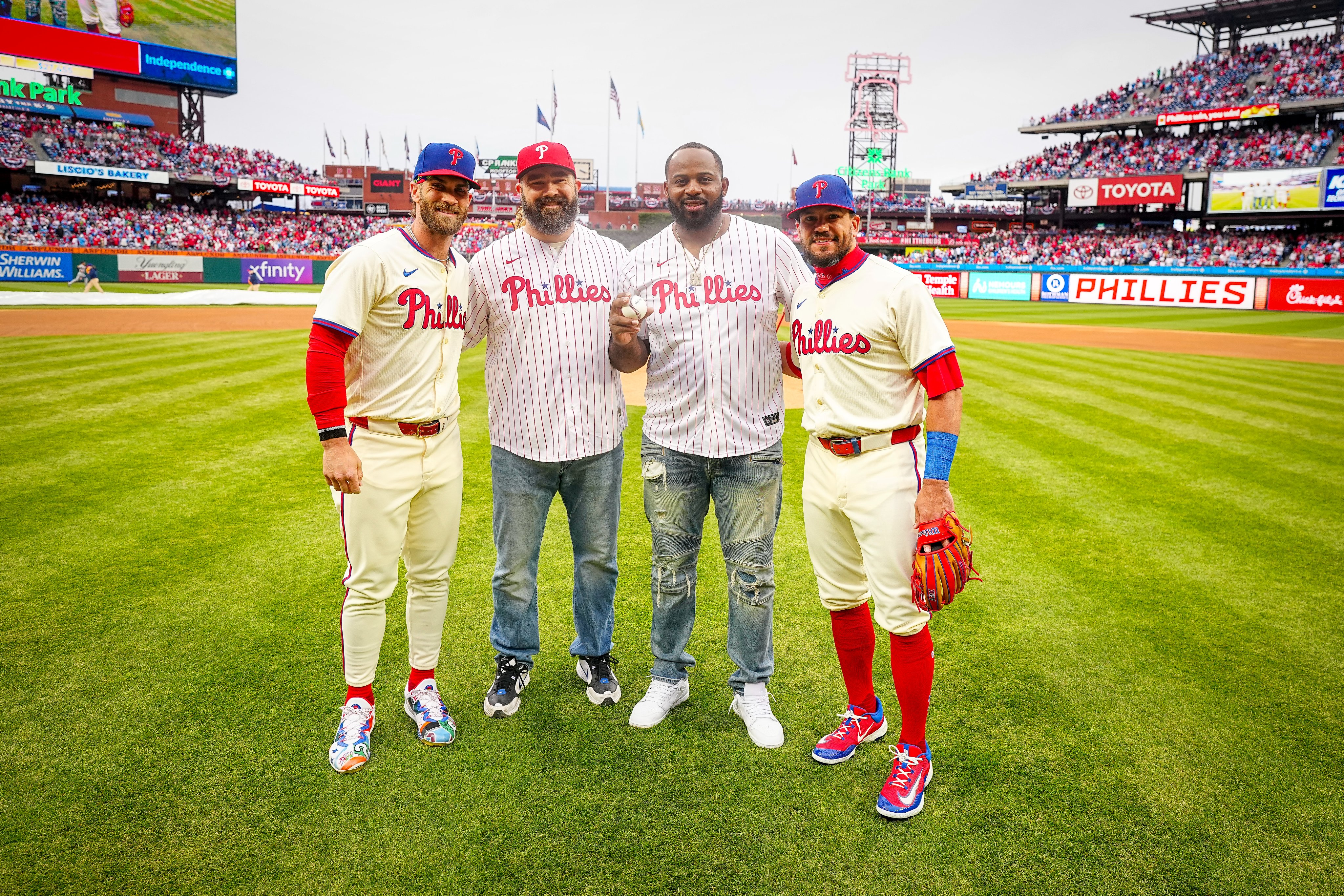 Bryce Harper, Jason Kelce, Fletcher Cox, and Kyle Schwarber all posing for a photo after Kelce and Cox threw out the first pitch. Harper and Schwarber are wearing the cream Phillies uniforms. Kelce and Cox are wearing the red pinstripes jerseys and jeans.