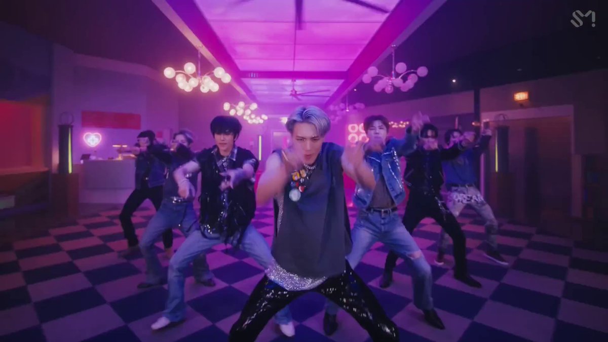 A group of male performers executes a synchronized dance routine in a purple-lit, checkered-floor room, with the SM Entertainment logo visible in the top right corner.