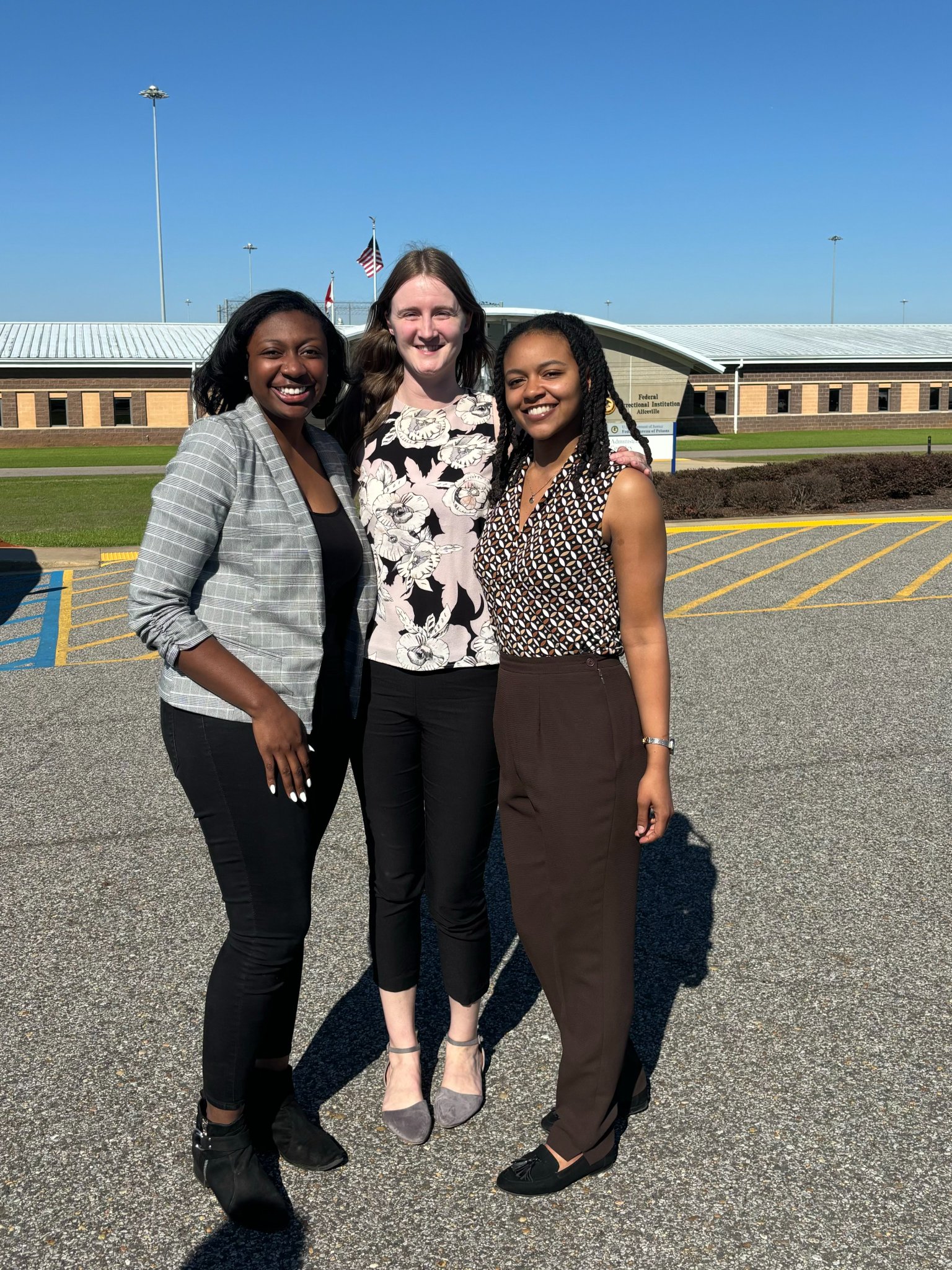 Three law students in a prison parking lot with a blue sky in the background above the prison. Aleah Brown is on the left, she is a Black woman wearing black slacks and a black and white blazer. Virginia Willis, in the middle, is a white woman wearing a black and white bouse and black slacks. Simone Hampton, on the right, is a Black woman wearing black slacks, a patterned blouse, and holding her blazer in her left hand. They are all smiling brightly. To protect her privacy, the client is not pictured.