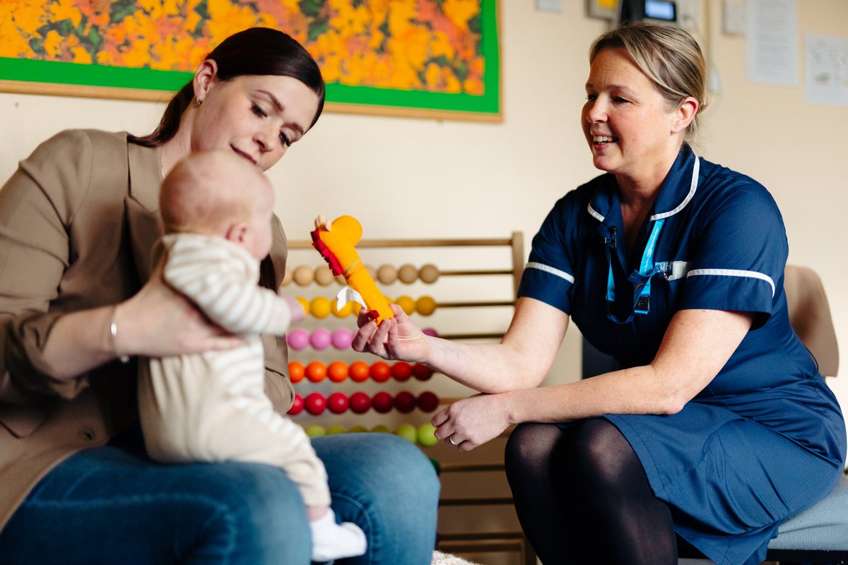 Health Visitor Rachel Rammage is holding up a toy for a baby to interact with. The baby is sat on its mother's lap.