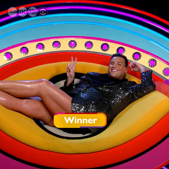 David in a black sequin jacket and short shorts, lying on the Diary Room chair doing a peace sign, with text saying ‘Winner’