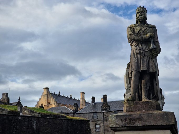 A stone statue of Robert The Bruce wearing armour and holding a sword. There is a Castle in the background.