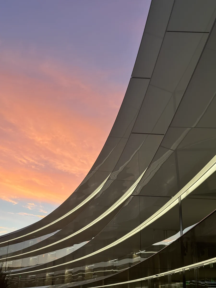 A photo of the ring building at Apple park during sunset. Architectural features of the building reflect Apple Park. The sky above is a gradient from blue to purple and the sun illuminates the clouds in a bright orange.