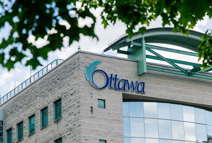 A close-up of Ottawa City Hall with the Ottawa logo sign centered. There are blurred leaves of a tree in the foreground. A sunny sky dotted with clouds is visible in the background.
