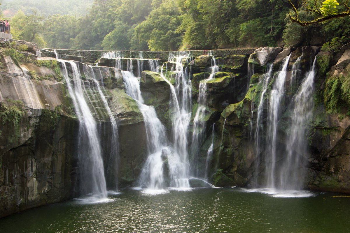 The Shifen waterfall is a wide waterfall cascading over rocky cliffs into a serene pond, surrounded by lush greenery with spectators on an observation deck to the left.

#throwbackthursday #taiwan #shifen #waterfall #WaterFeature #FlowingWater #Splashing #Tree #travel