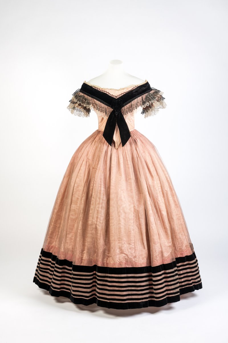 Friday Treat Time and we’re feeling pretty in pink with this oh so gorgeous evening dress from the early 1860s! Made from a beautiful silk moire, it features a fashionable low neckline that drops off-the-shoulder and is trimmed with a bertha style collar of black and white lace