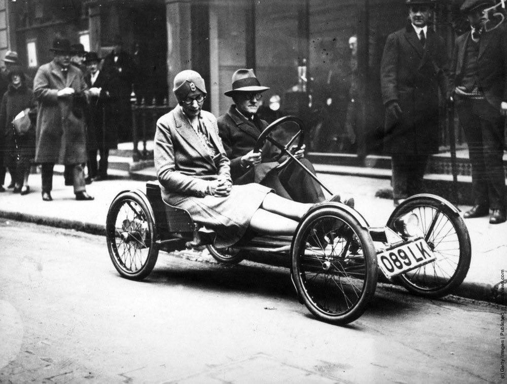 A man and a woman riding in an [Auto Red Bug], America’s latest electric 2-seater runabout, in a London street.  Driven by a 16-volt battery, it is capable of 12 mph. 1929.

The Auto Red Bug, also known as the Smith Flyer, is an early example of a microcar or what some might