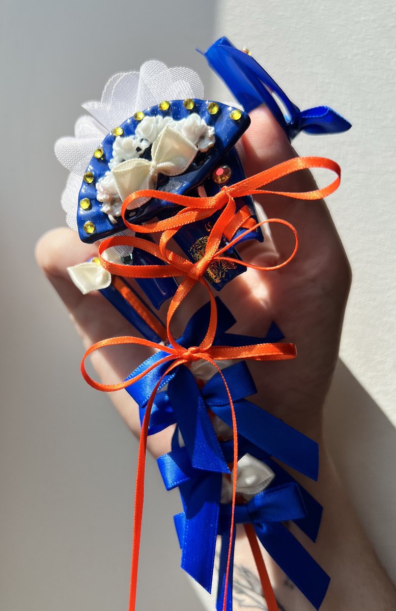 picture of a hand with intricate royal blue square nail art that has a small sculpted fan, with rhinestones, and many blue, white and orange ribbons tied into bows.