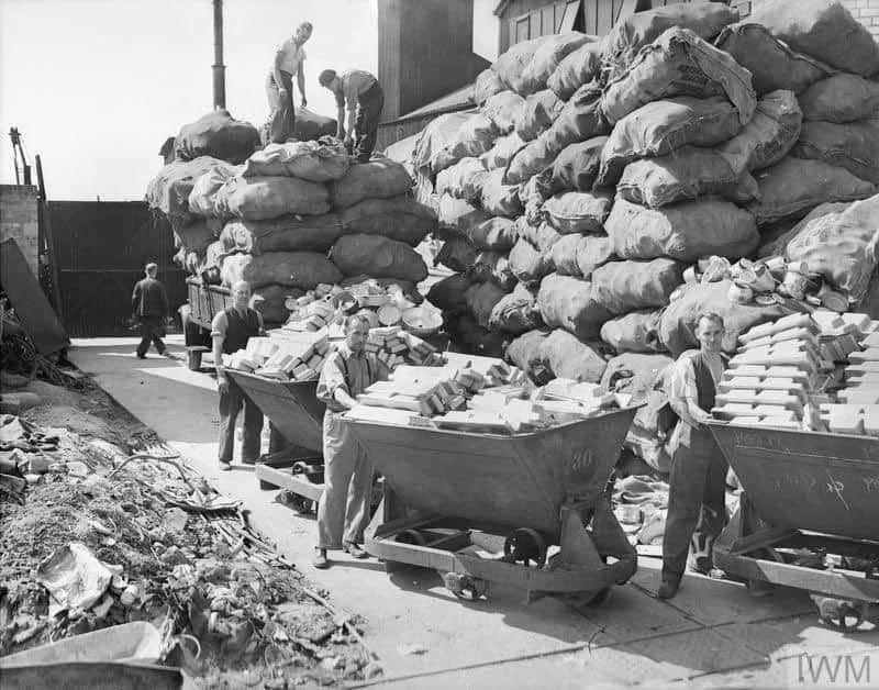 A photograph of Metal workers wheeling large barrows full of ingots of pure aluminium from a workshop, somewhere in Britain. 
In the background, huge piles of sacks of aluminium items can be seen, from which workers select further items for smelting.