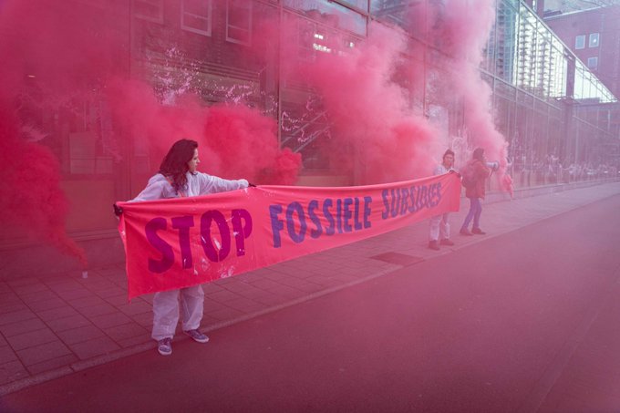 2 rebellen houden een banner vast met de tekst Stop fossiele subsidies. Rechts staat een derde rebel met een megafoon. Ze staan voor het ministerie van Economische Zaken en klimaat, waar roze verf op de ramen is gesproeid. De rebellen worden omgeven door roze rookwolken. 