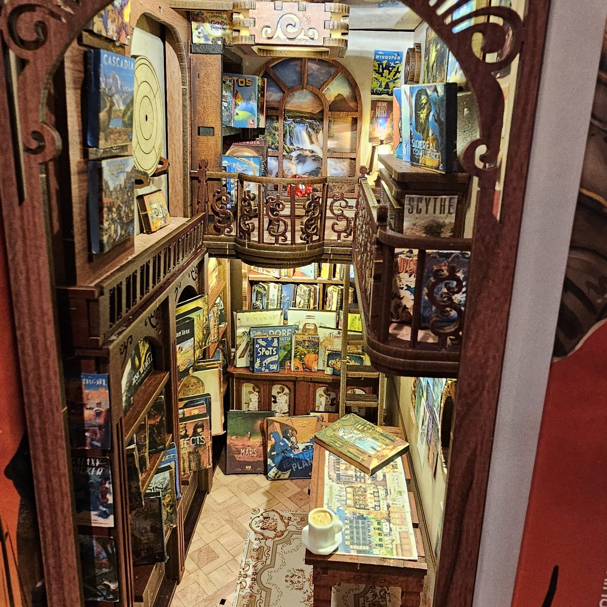time_of_corvid's tweet photo. I made not a book nook but a boardgame nook, tucked gently between @garphillgames boxes. Weather Machine by @vitallacerda and @ianotooletweets is on the table but I'd NEVER put a cup of tea on it in real life. https://t.co/r88IlvXr9w