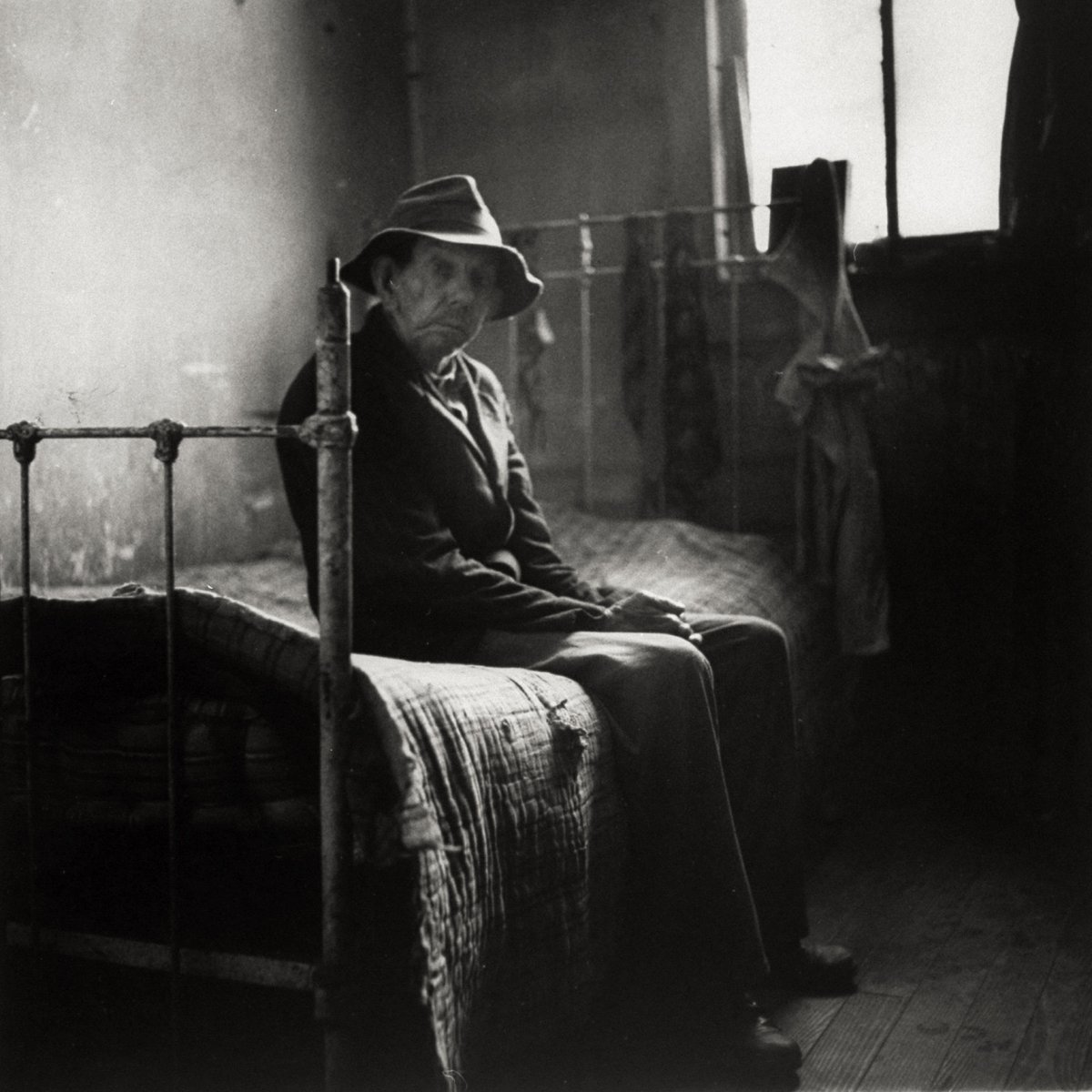 An old man sitting patiently on an old iron bed as he waits for mealtime at a poorhouse, Perry County, Pennsylvania, ca. 1952 - by Wallace Kirkland (1890 - 1983), American