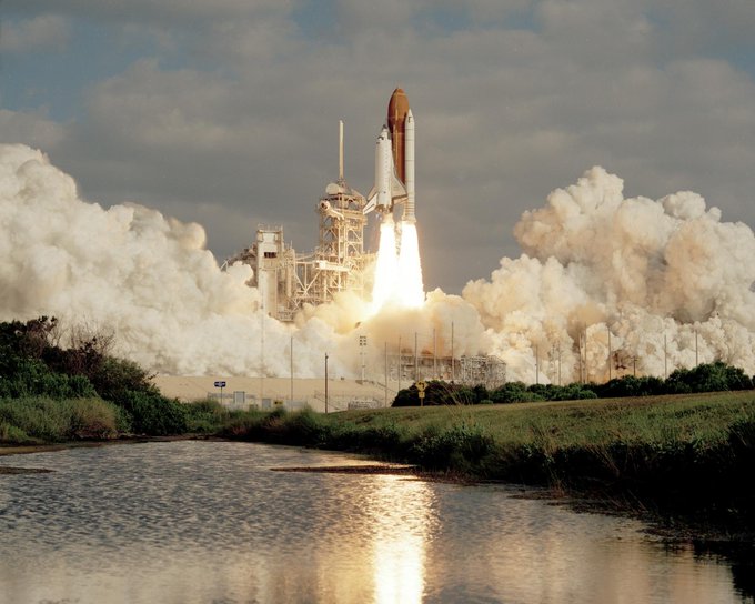 A space shuttle launching while attached to a rocket. At the bottom of the image there is a lake reflecting the launch while smoke fills the areas above.