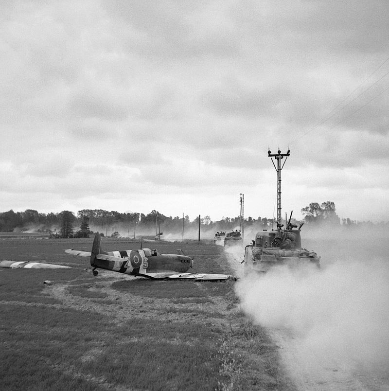 Sherman tanks move up past a crash-landed Spitfire, for an attack on Tilly-sur-Seulles, 17 June 1944. The Spitfire Mk IXb squadron code – 'VZ' – indicates it belongs to RCAF 412 Squadron.