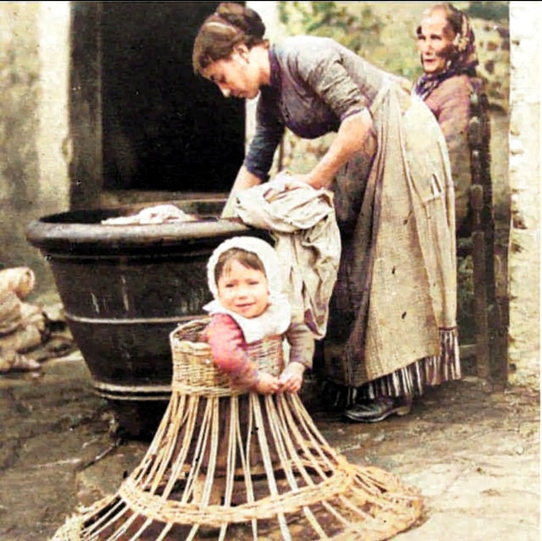 A toddler learning how to walk in a wicker frame while mum gets on with the washing, 1910. #edwardians #edwardianperiod #parentinglife #colourised