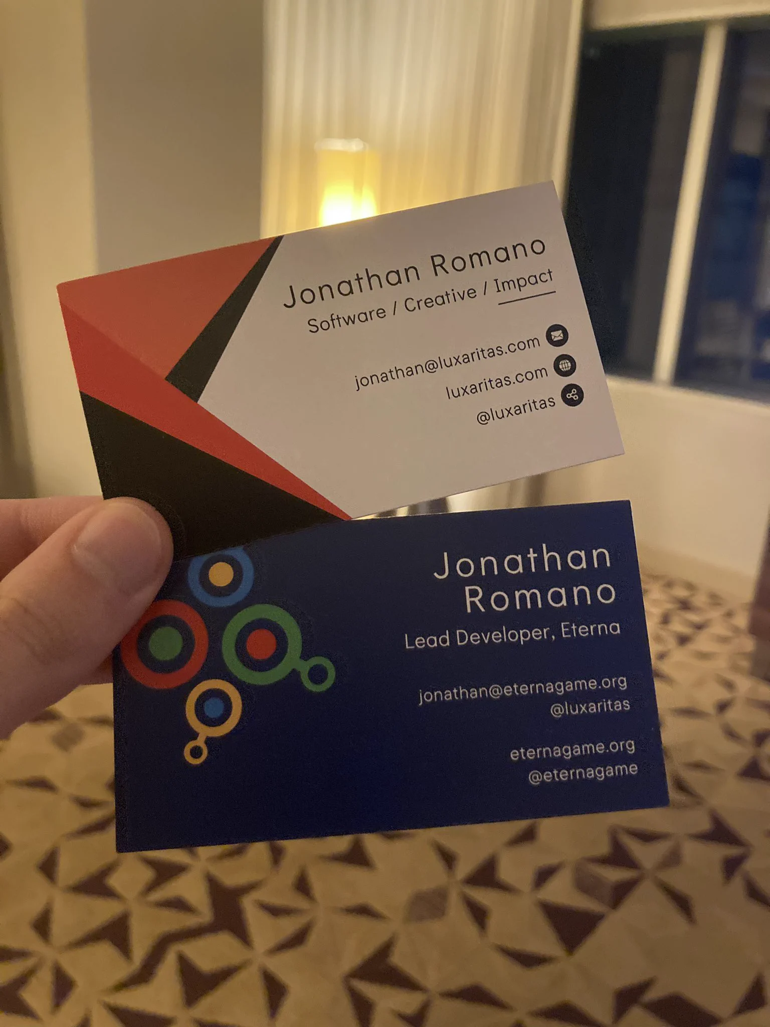 Two business cards, one on the bottom branded with the Eterna logo of a combination of blue, yellow, green, and red circles, titled “Lead Developer, Eterna”, and one on the top with angular red and black triangles, titled “Software / Creative / Impact”; both contain various contact details for myself