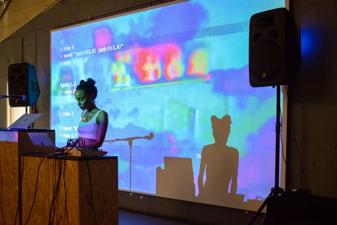 Performer standing behind a wooden platform with two laptops, speakers and lots of wires. They are illuminated by a large blue/pink/purple projection of computer code.