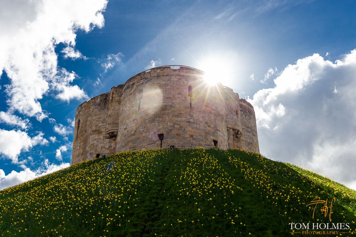"Clifford The Big Stone Tower"
The imposing 'Clifford's Tower' in York (North Yorkshire, UK). 
The bright sunshine, fluffy clouds and daffodils contrast the buildings varied and dark history.

© Tom Holmes / buff.ly/2q2s0de