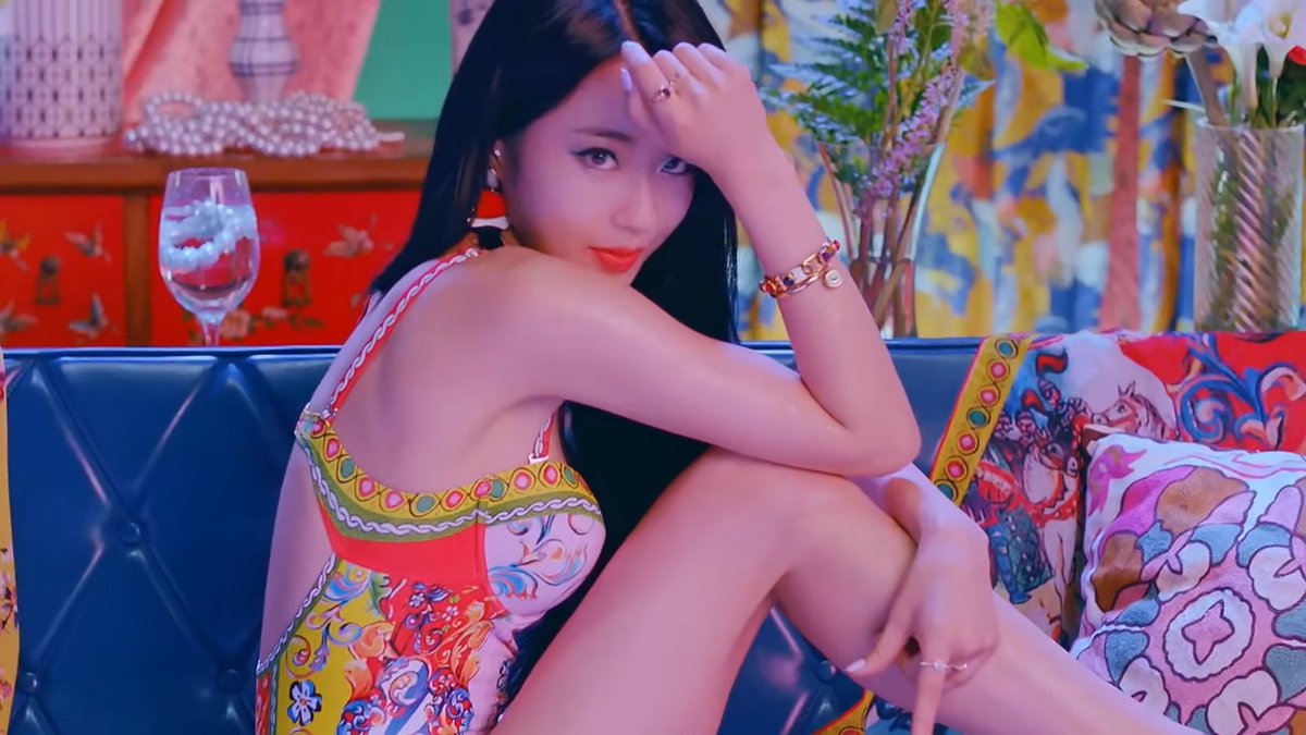 A young woman posing on a blue couch in a vibrantly decorated room, wearing a colorful patterned outfit with red lipstick, bracelets, and a ring.