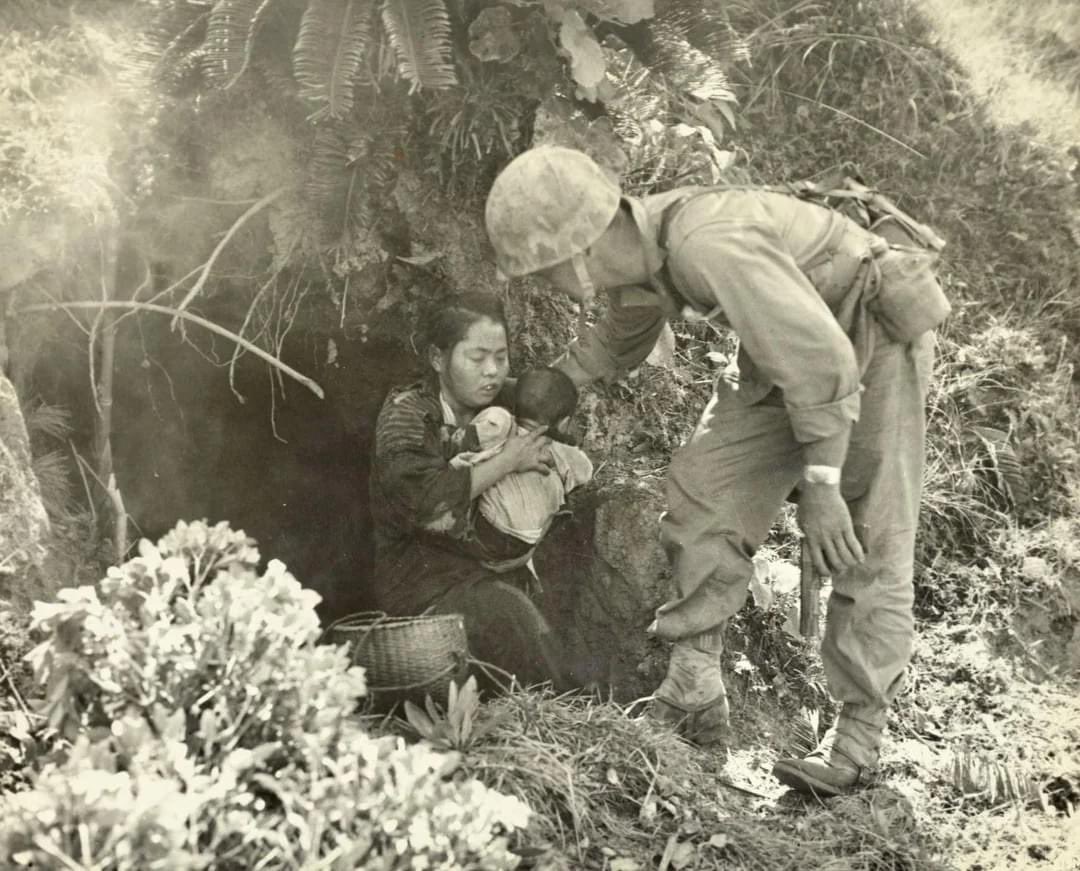 Eleven Okinawa civilians who were huddled in this hillside cave were rescued when a passing Marine patrol heard a baby crying. After being assured that no harm would come to them they emerged from their hideout and here a Leatherneck lends a hand to a mother and baby, 1945.