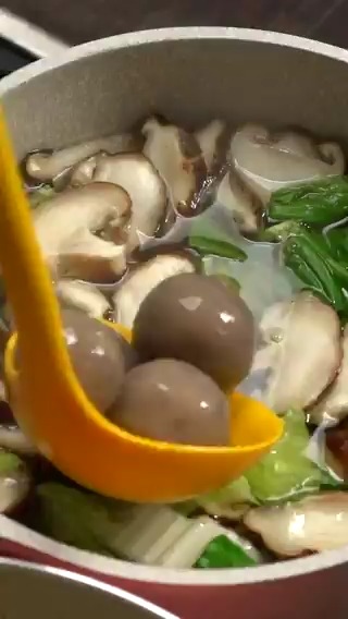 A close-up of a pot of soup with mushrooms, leafy greens, and noodles, with a yellow ladle scooping up what appears to be quail eggs or meatballs.